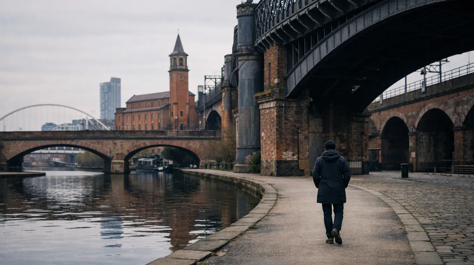 A man walking alone along Manchester's canalside beneath the railway viaducts on a grey day