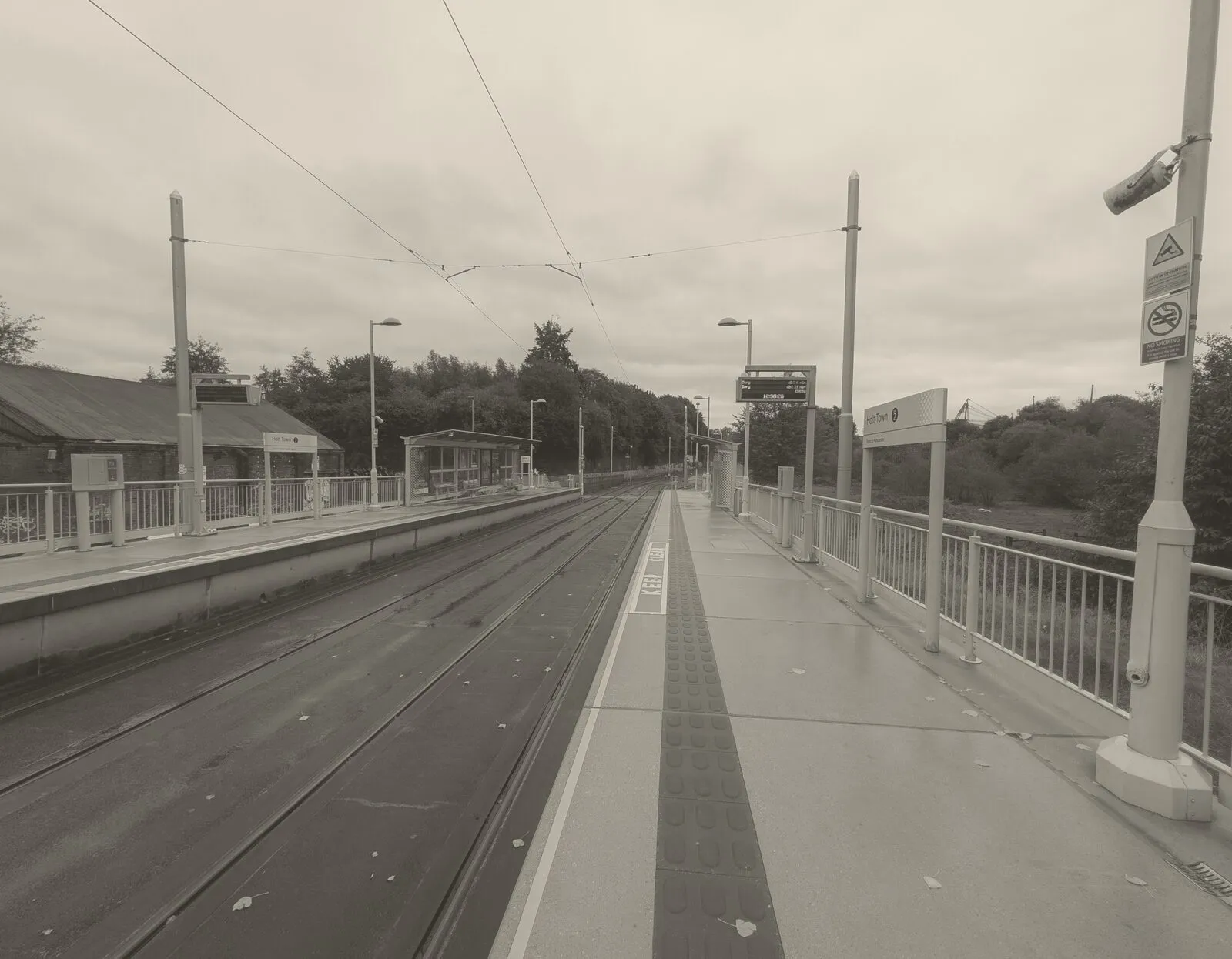 An empty tram platform at Holt Town on a wet morning, the tracks running away into the distance