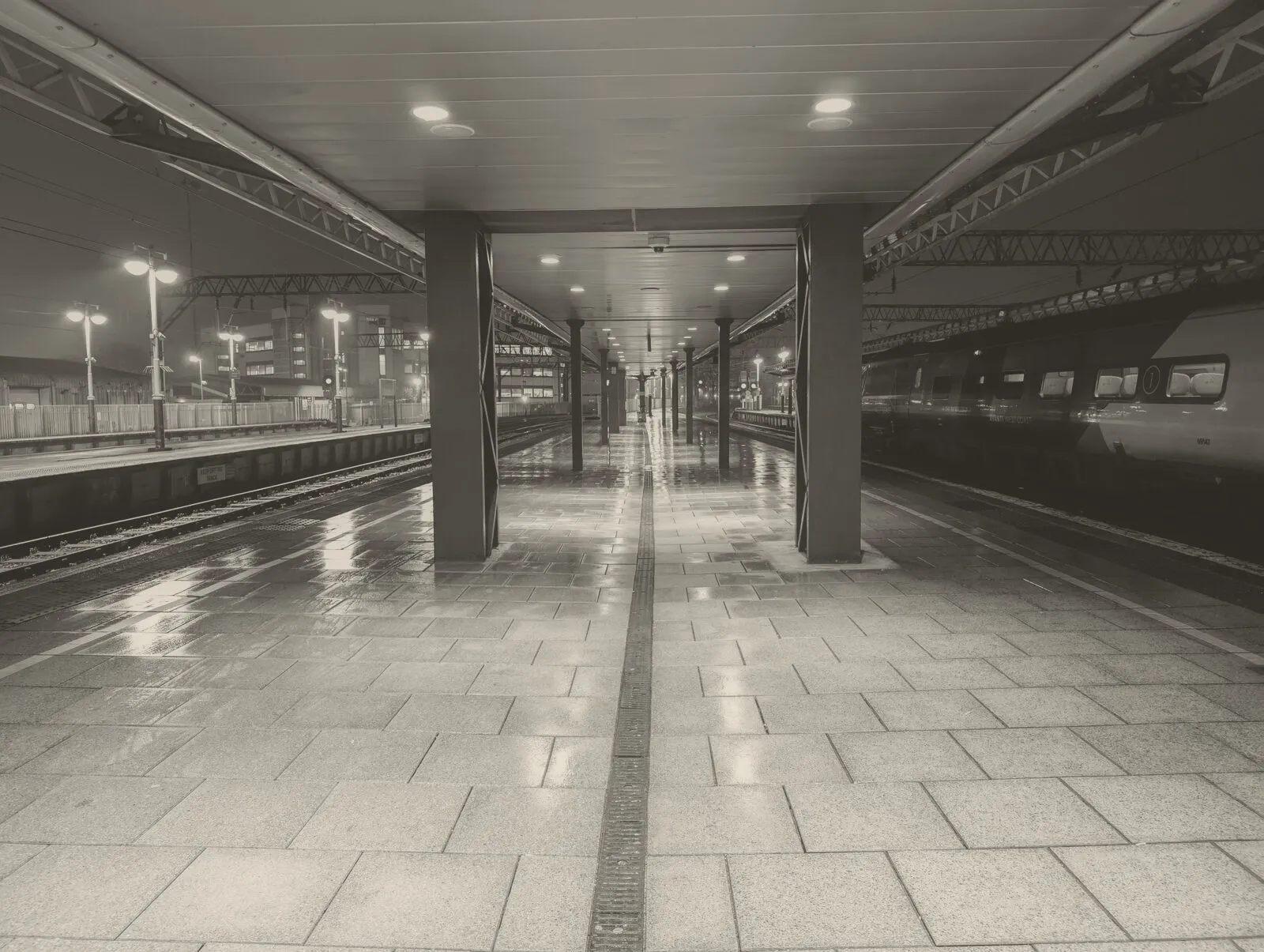 A wet, empty platform at Manchester Piccadilly at night, the lights reflected in the paving