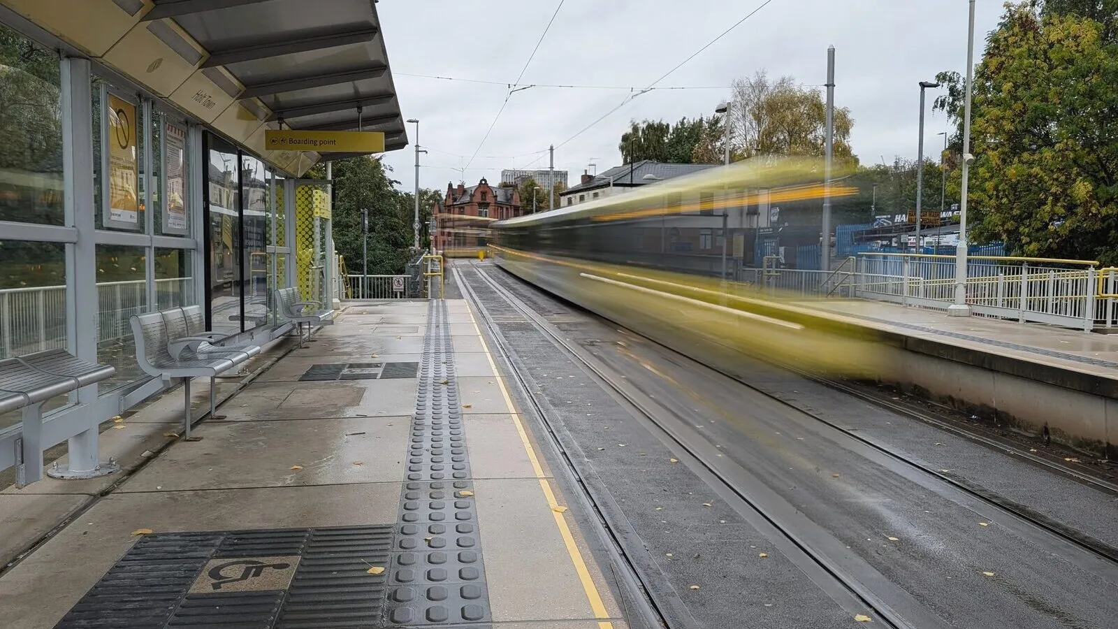 A Metrolink tram blurring past an empty platform at Holt Town on a grey Manchester morning